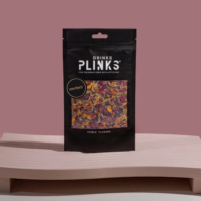 Black package of Drinks Plinks confetti edible flowers on a wooden surface with a pinkish-brown background