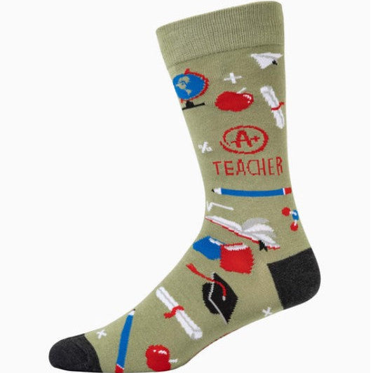 An olive green crew sock with a playful 'Teacher' theme, featuring school-themed illustrations like a globe, apple, book, and graduation cap, along with the text 'A+ TEACHER' and black heels/toe caps.