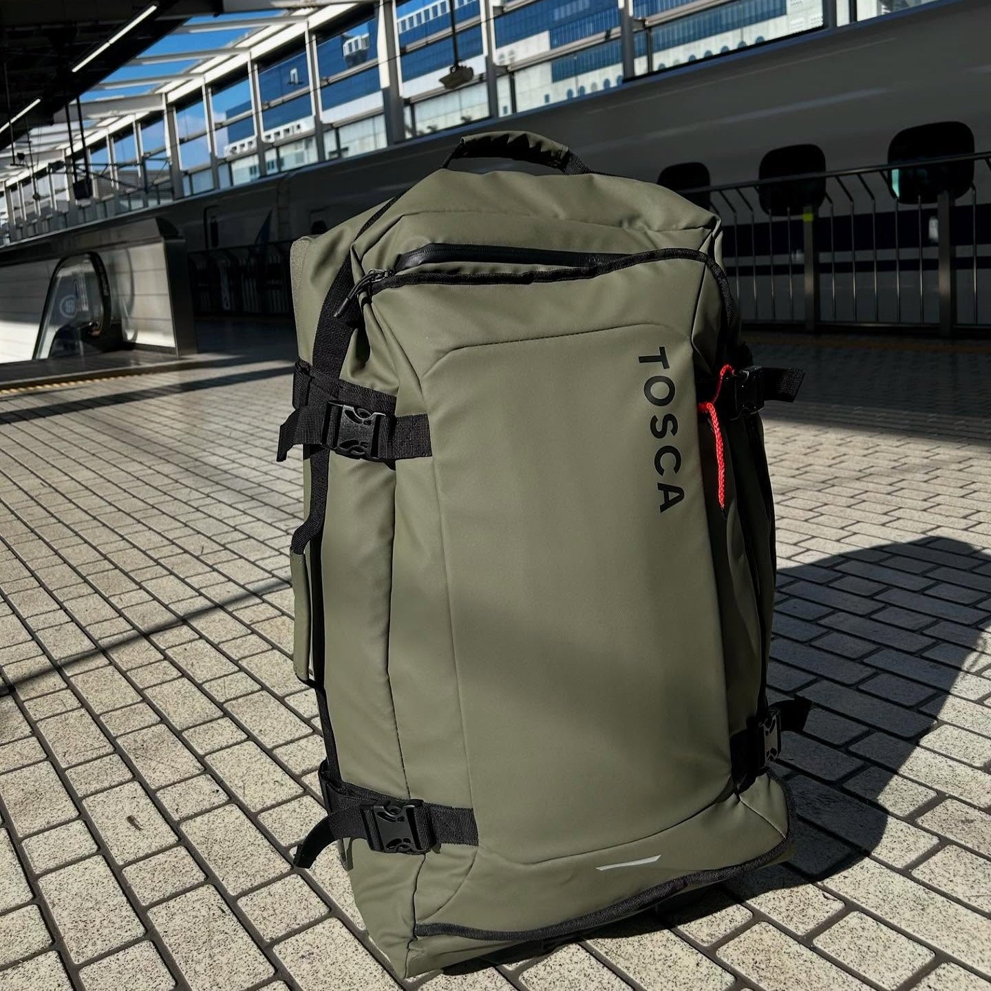 Green backpack with 'TOSCA' branding on a pavement near a train station.