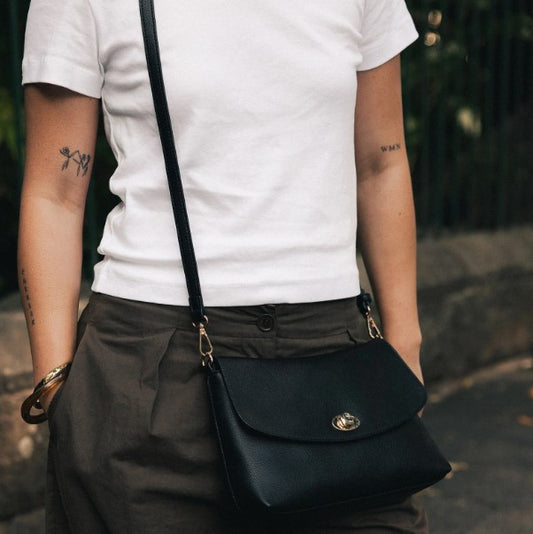 A person wearing a white t-shirt and dark olive pants carries a black Louenhide crossbody bag with gold-tone hardware and a turn-lock closure.