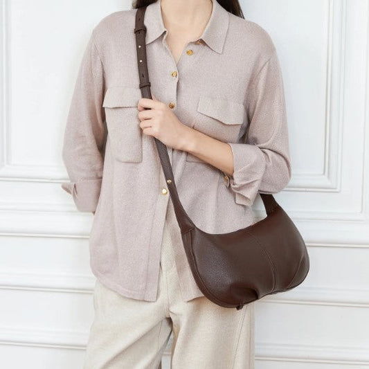 Person wearing a light gray shirt and beige pants with a brown leather shoulder bag against a white wall.