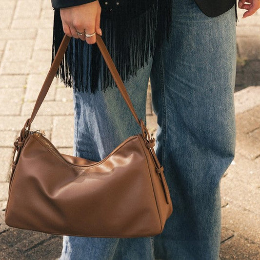A person wearing blue jeans and a black fringed top carries a brown shoulder bag on a paved outdoor walkway.