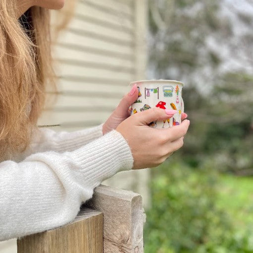 A person in a white knit sweater holds a small paper cup with a colorful illustrated pattern outdoors, leaning on a wooden railing with a blurred house and greenery in the background.