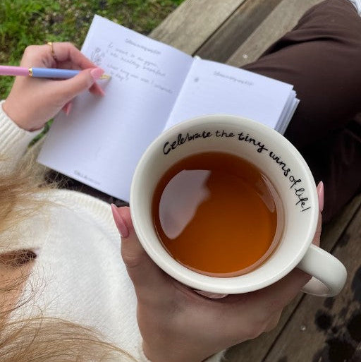 A person holding a white ceramic mug filled with tea, with the phrase 'Celebrate the tiny wins of life!' printed in black script inside the mug. Another hand holds an open notebook and a pastel pen, and a person's legs are visible in the background on a wooden bench.