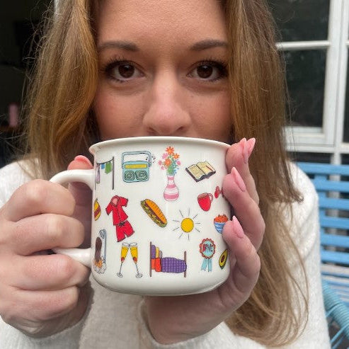 A woman holds a white ceramic mug with a colorful retro-themed illustration pattern, featuring icons like a cassette tape, flowers, a book, and a sun, in front of her face.