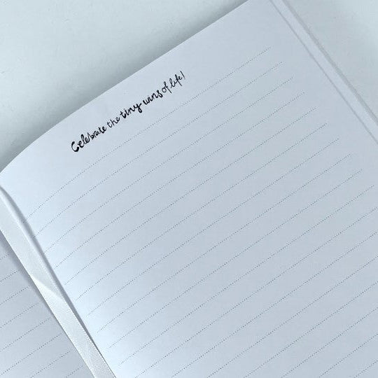 A close-up of an open lined notebook page with a handwritten note reading 'Celebrate the tiny wins of life!' in black ink, photographed on a clean white background.