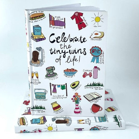 A white hardcover journal with colorful hand-drawn doodles and the phrase 'Celebrate the tiny wins of life!' on the cover, shown standing upright with a matching wrapped gift or folded journal on top against a clean white background.