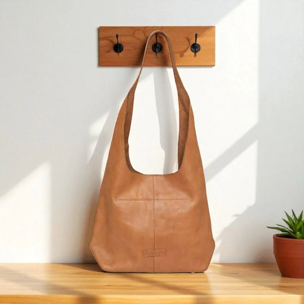 Brown leather bag hanging on a wooden hook against a white wall with a plant in the corner.