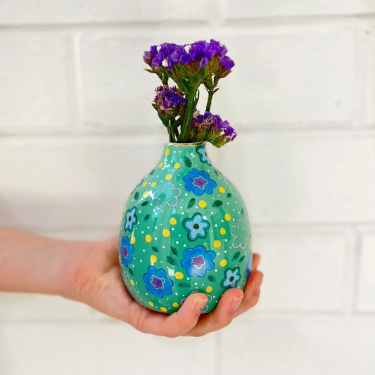 Hand holding a green vase with floral patterns and purple flowers against a white tiled wall.