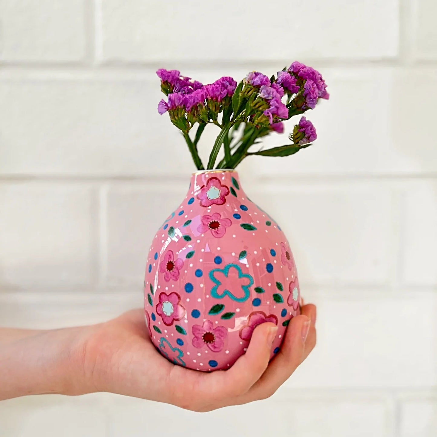 Hand holding a pink decorative vase with floral patterns and purple flowers against a white tiled wall.
