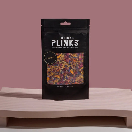Black package of Drinks Plinks confetti edible flowers on a wooden surface with a pinkish-brown background