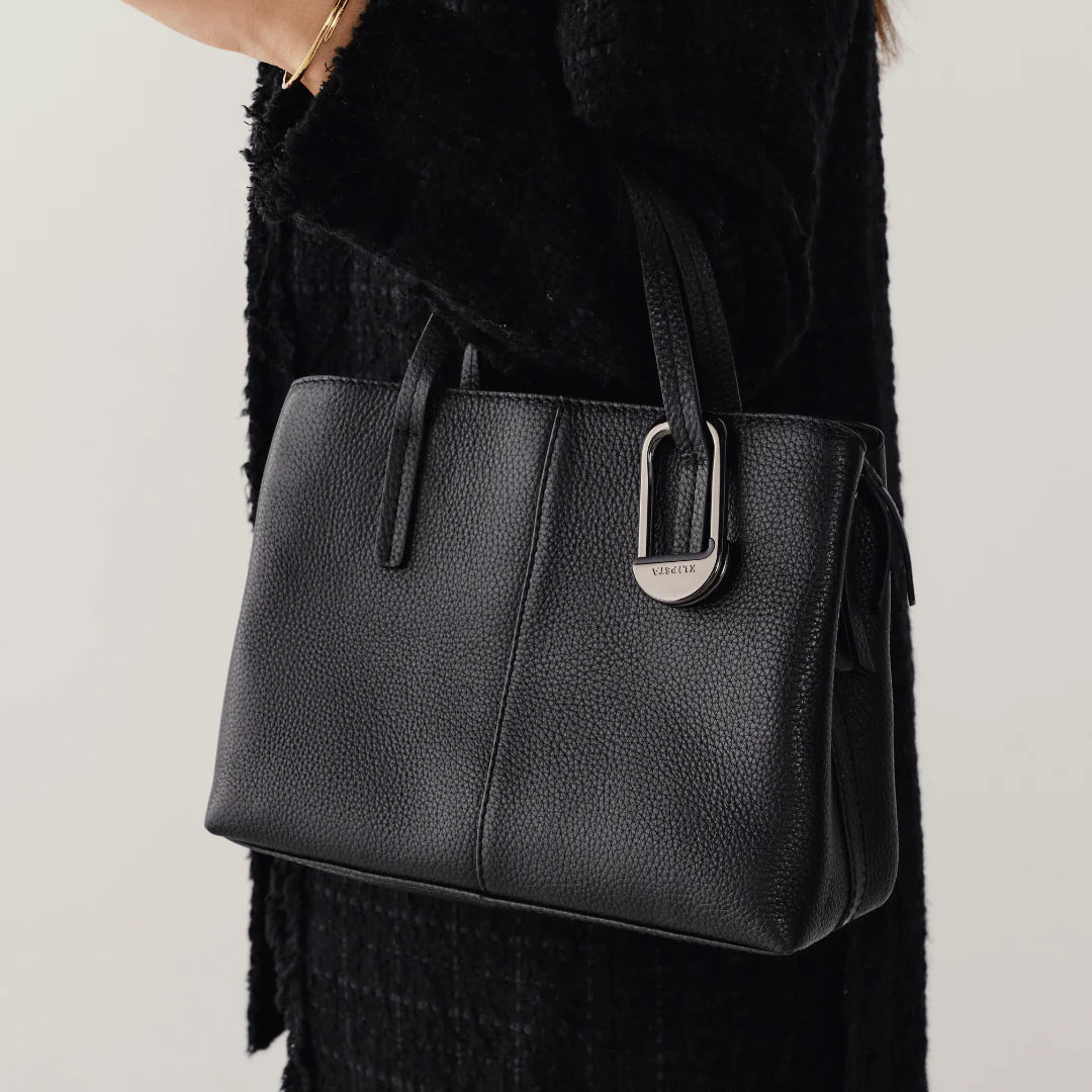 Black leather handbag with a brand logo held by a person wearing a black coat.