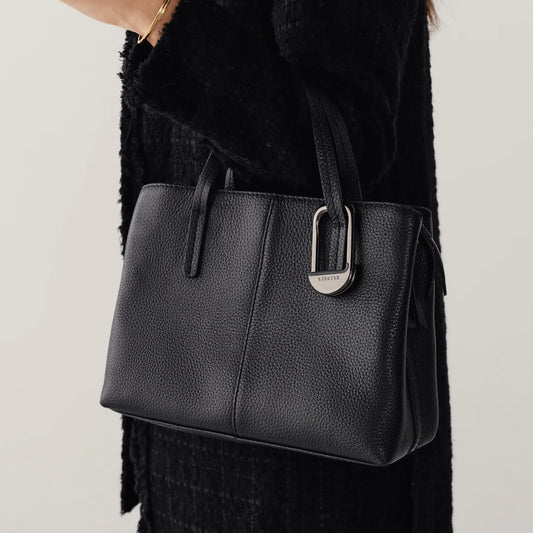 Black leather handbag with a brand logo held by a person wearing a black coat.