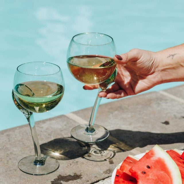 Two wine glasses with a hand holding one, next to a slice of watermelon on a stone surface with a blue sky background.