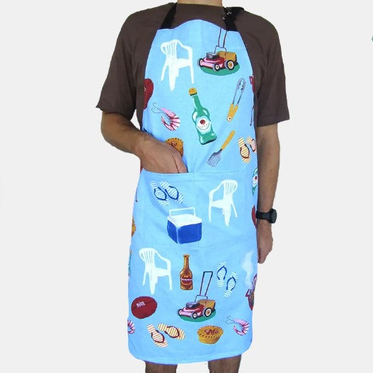 A person wearing a light blue apron with a brown short-sleeve shirt underneath. The apron features a colorful all-over print of summer and backyard items like white plastic chairs, a lawnmower, a beer bottle, a cooler, and a whistle, with a front pocket.
