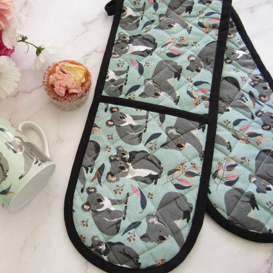 Oven mitts with koala design on a marble surface with a mug and flowers.