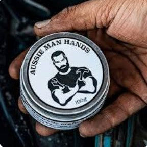 A hand holding a round tin of Aussie Man Hands hand cream, with the label clearly showing the brand name and a graphic of a bearded man with flexed arms.