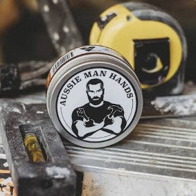 A round metal tin labeled 'AUSSIE MAN HANDS' with a black-and-white illustrated figure, photographed on a workbench with tools and a yellow tape measure in the background.
