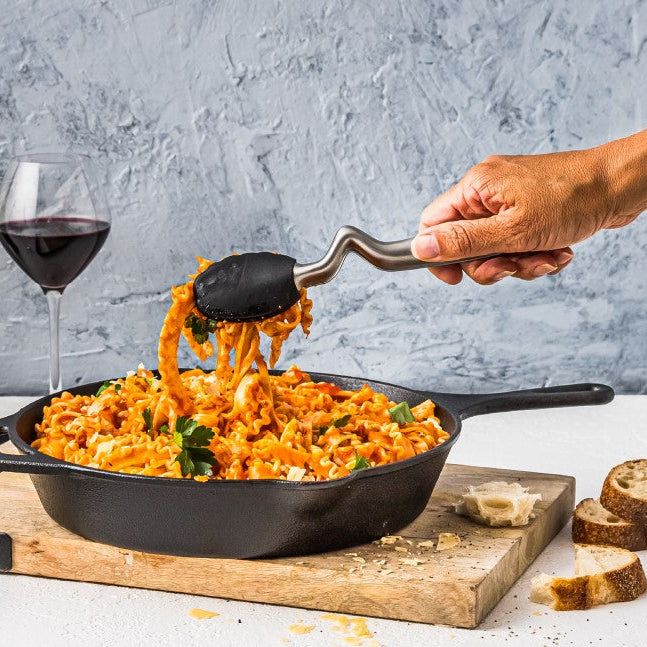 A cast iron skillet filled with orange-red pasta on a wooden cutting board, with a hand using a utensil to lift the pasta. A glass of red wine and slices of bread are visible against a textured gray background.