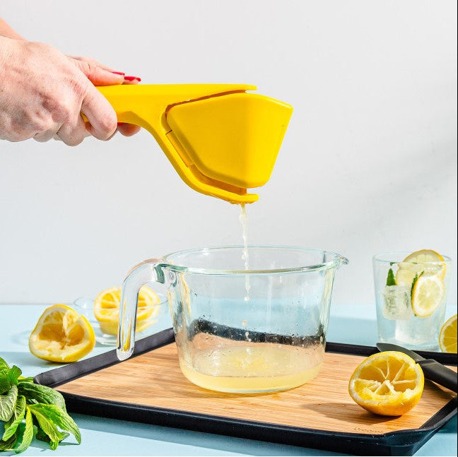 A hand uses a bright yellow handheld citrus juicer to squeeze juice into a clear glass measuring cup. Lemons, mint leaves, and a glass of lemonade are arranged on a wooden board against a clean white background.