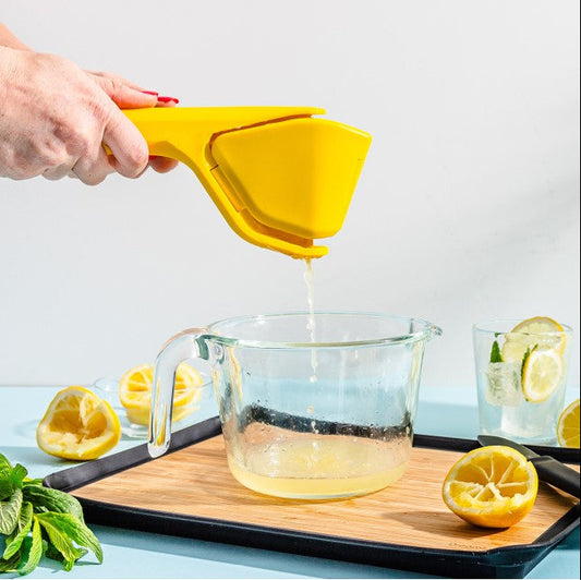 A hand uses a bright yellow handheld citrus juicer to squeeze juice into a clear glass measuring cup. Lemons, mint leaves, and a glass of lemonade are arranged on a wooden board against a clean white background.