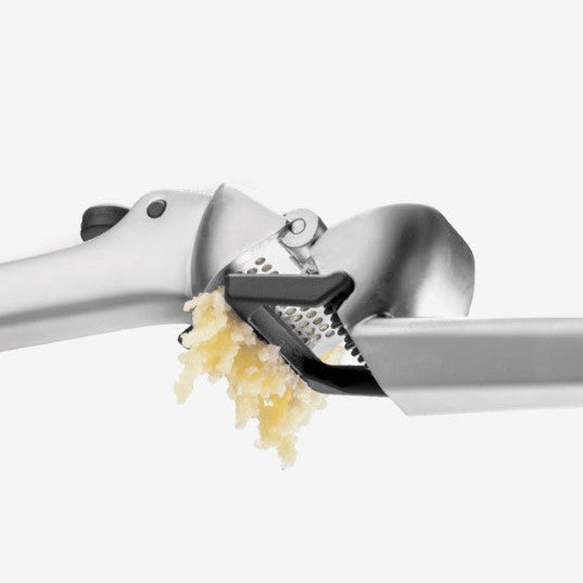 A close-up of a stainless steel garlic press with a black handle, shown pressing garlic cloves that are falling out, on a clean white background.