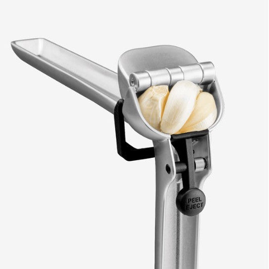 A stainless steel garlic press with peeled garlic cloves inside, shown on a light gray background.