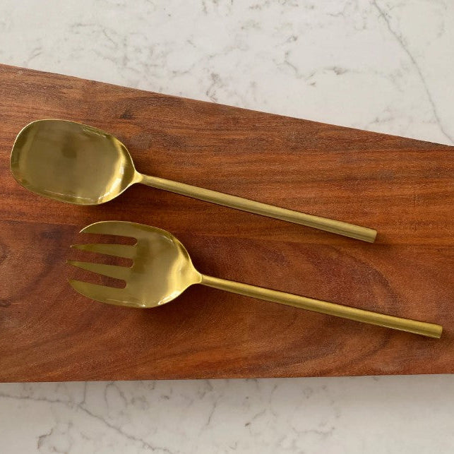 A gold-tone metal serving spoon and matching serving fork resting on a rectangular wooden board over a white marble surface.