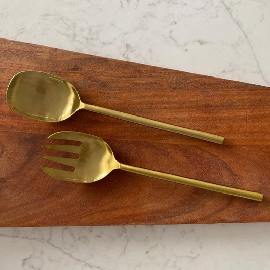 A gold-tone metal serving spoon and matching serving fork resting on a rectangular wooden board over a white marble surface.