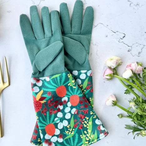Pair of green gardening gloves with colorful floral pattern on a white marble surface with flowers and a gold tool.