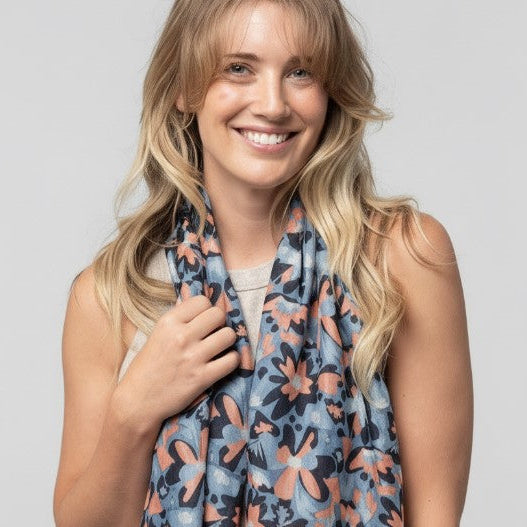 A smiling blonde woman wearing a lightweight floral scarf in muted blue, peach, and gray tones, photographed against a plain light gray studio background.