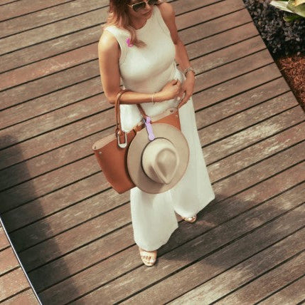 Woman in a white dress holding a brown handbag and a straw hat on a wooden deck.