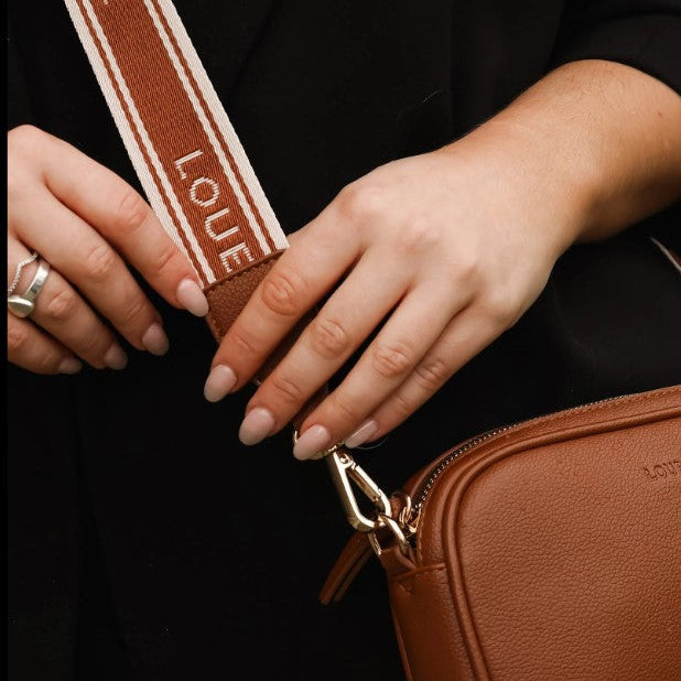 Brown leather bag with a strap labeled 'LOUENHIDE' held by hands against a dark background