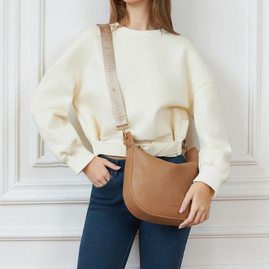 Person wearing a beige sweater and blue jeans holding a brown leather bag against a white wall.