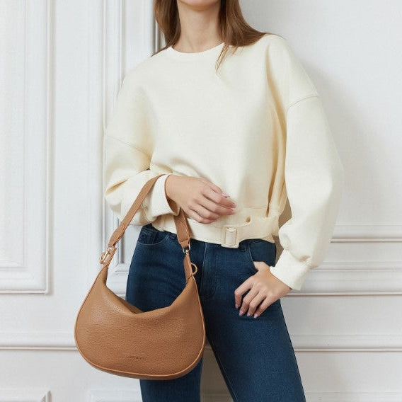 Person wearing a cream sweater and blue jeans holding a brown handbag against a white wall.