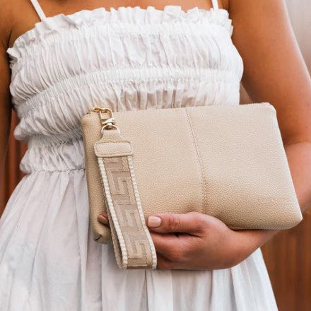 Beige clutch bag with a strap held by a person wearing a white dress.