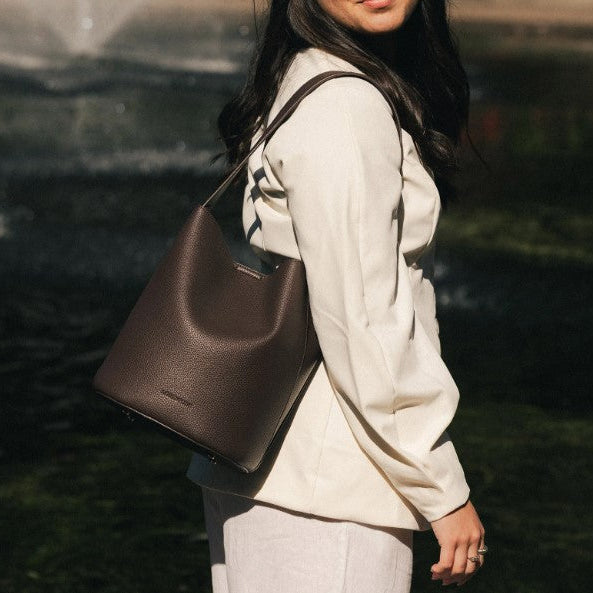A woman in a cream-colored outfit carries a dark brown pebbled bucket bag over her shoulder, set against a blurred outdoor background.