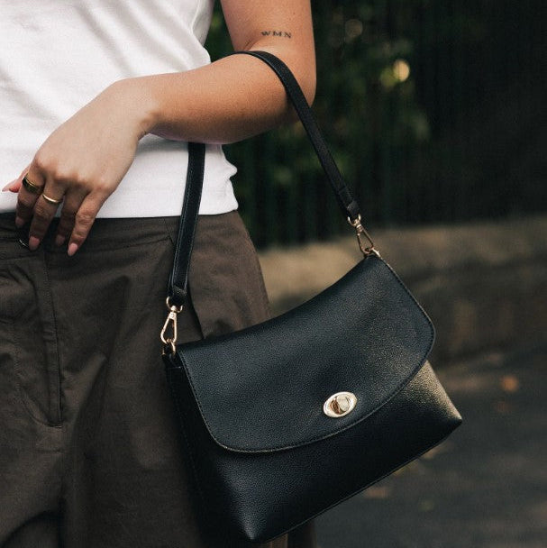 A person wearing a white t-shirt and brown pants carries a black Louenhide shoulder bag with a gold-tone turn-lock closure in an outdoor setting.