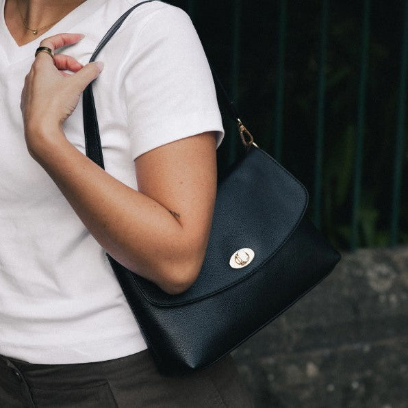 A person wearing a white t-shirt and dark pants carries a black Louenhide shoulder bag with a gold-tone turn-lock closure and gold hardware.