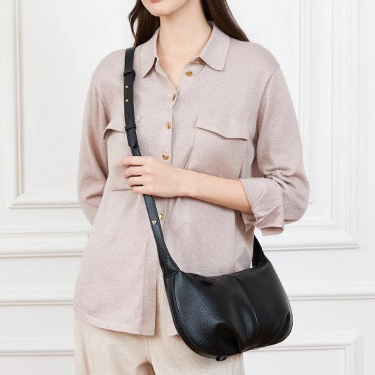 Person wearing a beige shirt and holding a black leather bag against a white wall.
