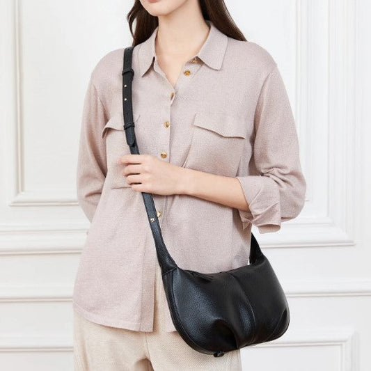 Person wearing a beige shirt and holding a black leather bag against a white wall.