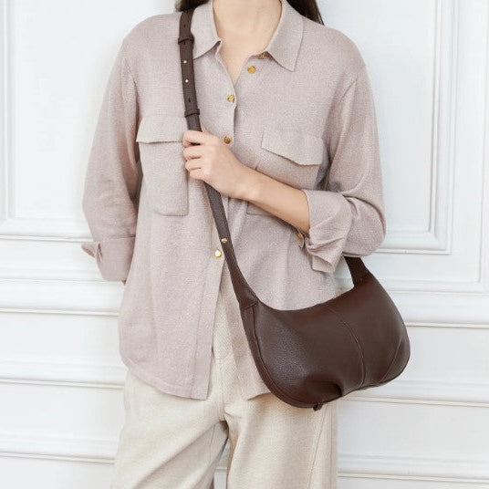 Person wearing a light gray shirt and beige pants with a brown leather shoulder bag against a white wall.