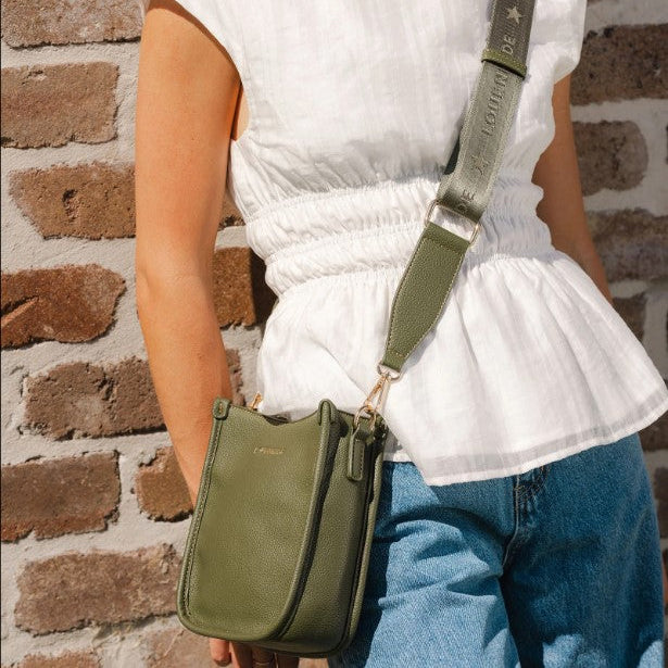 Person wearing a white top and blue jeans holding a green handbag against a brick wall.