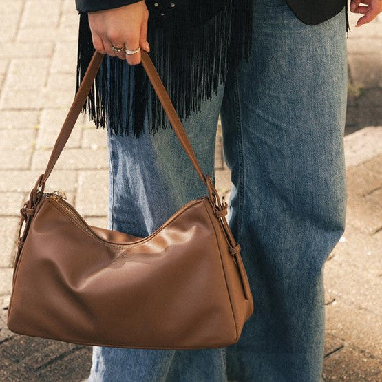A person wearing blue jeans and a black fringed top carries a brown shoulder bag on a paved outdoor walkway.