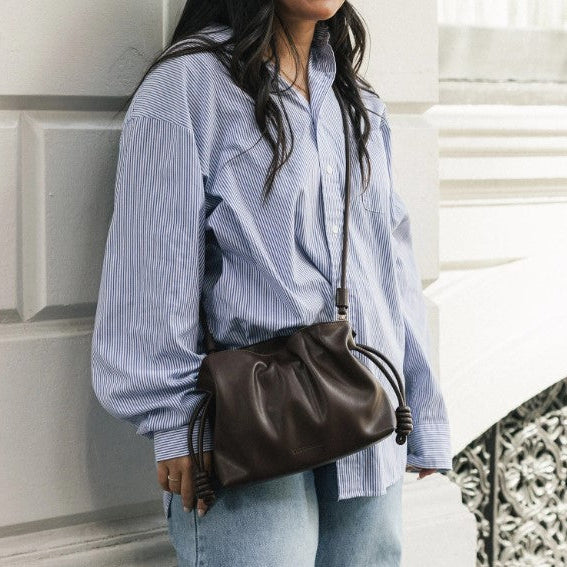 A woman wearing a blue and white striped button-up shirt and light wash jeans carries a dark brown crossbody bag with a drawstring detail, standing against a white paneled wall.