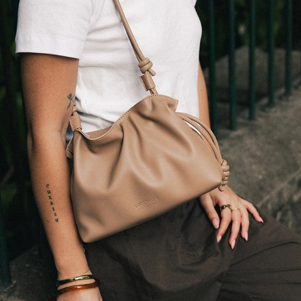 A person wearing a white t-shirt and dark brown pants carries a beige  shoulder bag with a knotted strap detail. The brand name 'LOUENHIDE' is subtly embossed on the front of the bag.
