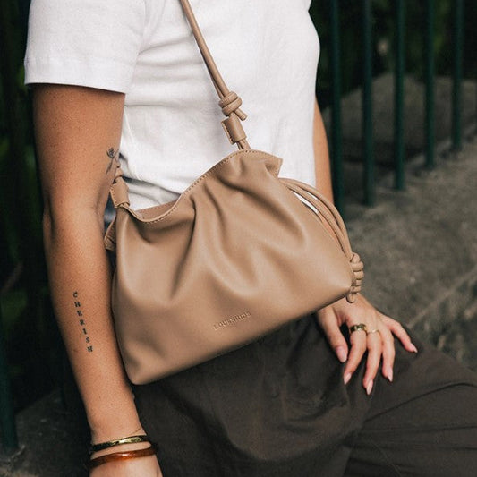 A person wearing a white t-shirt and dark brown pants carries a beige  shoulder bag with a knotted strap detail. The brand name 'LOUENHIDE' is subtly embossed on the front of the bag.