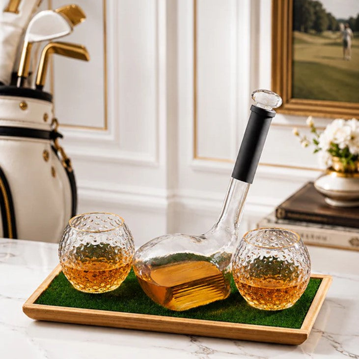 A clear glass decanter with a black handle and two matching textured glass tumblers sit on a green tray in a styled interior setting with a golf bag and framed picture in the background.