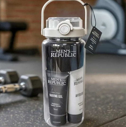 A clear cylindrical travel shower bottle with a white handle and lid, shown on a gym floor with dumbbells and a bench in the blurred background. The bottle contains Men's Republic shaving gel and an after-shave tube, with a black hangtag attached.