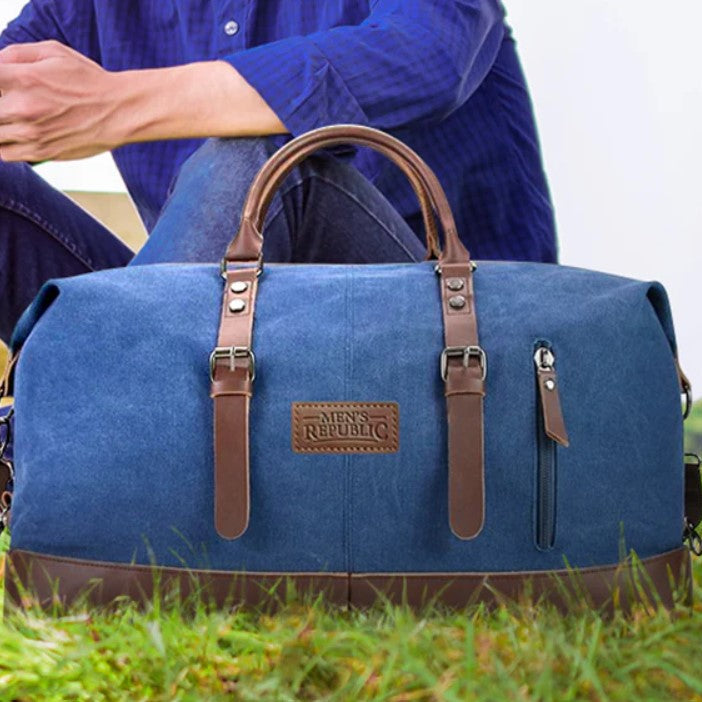 Blue duffel bag with brown straps on grass, person wearing blue shirt in background